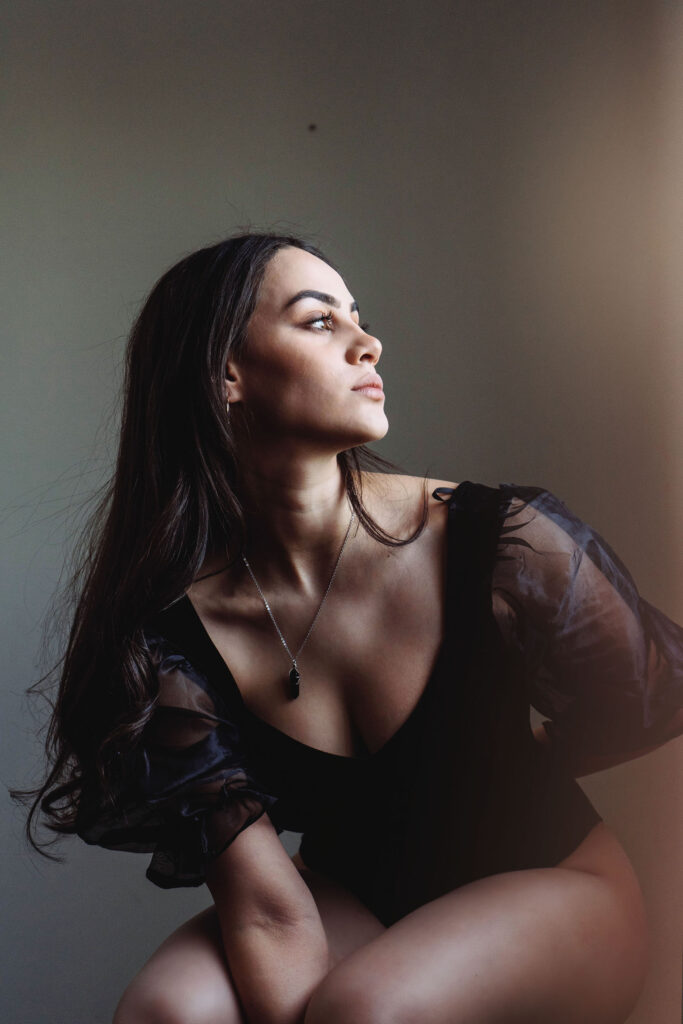 Valeria Cisneros a model with long dark hair is captured in a side profile gazing upwards. She wears a black outfit with sheer sleeves and a necklace with a pendant. The lighting casts a soft glow on her face emphasizing her features.